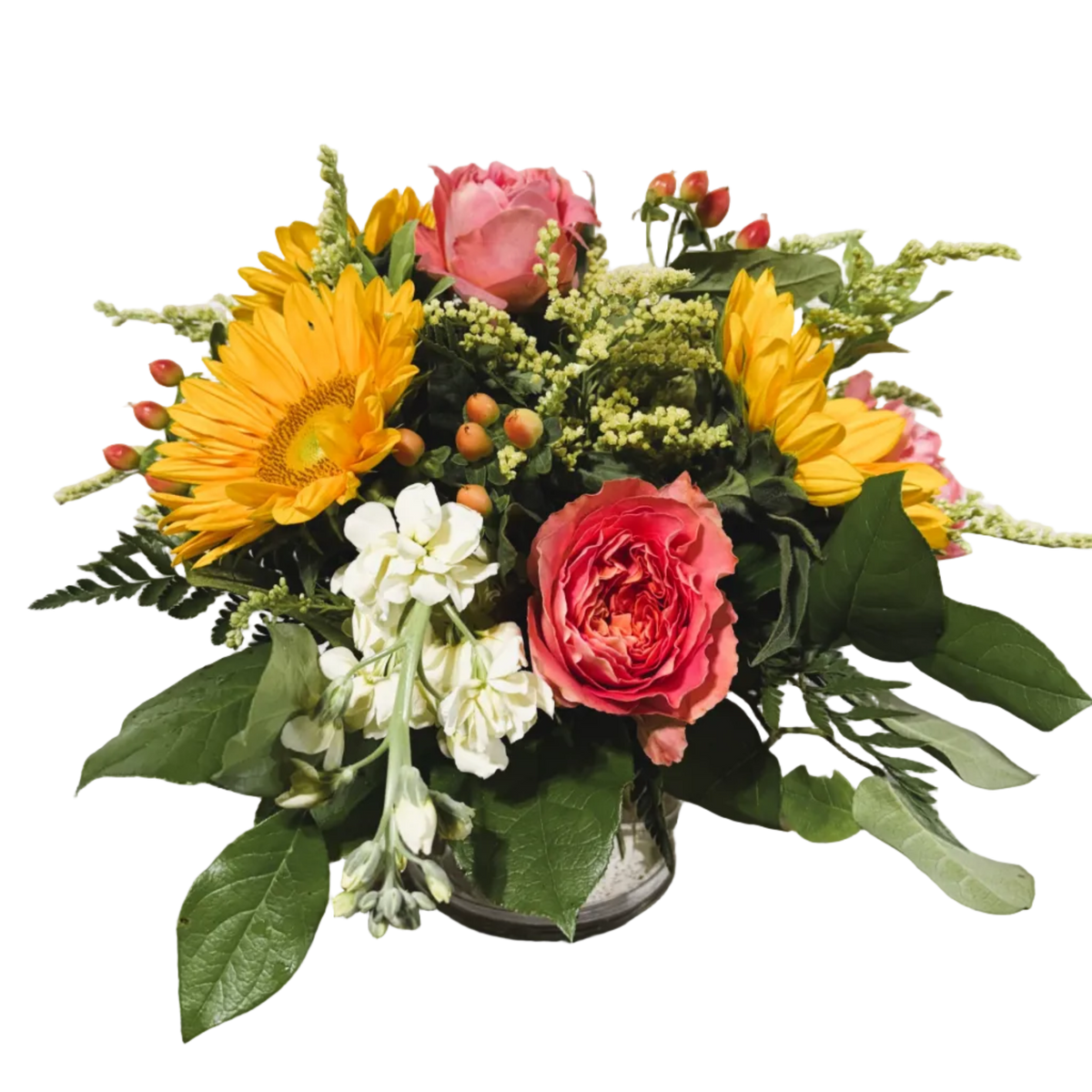 A vibrant flower arrangement in a small clear glass vase, featuring yellow sunflowers, pink roses, cream stock, and green foliage on a solid white background.