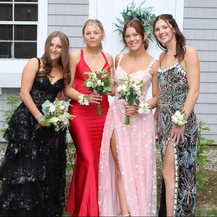 Four women in formal dresses holding flowers in front of a house.