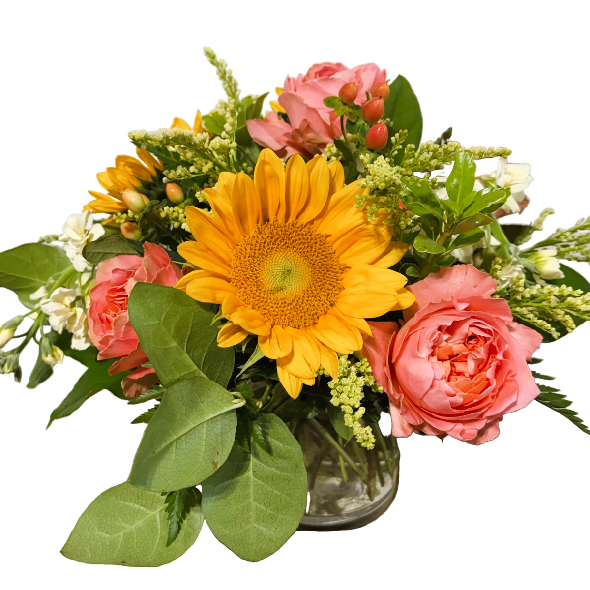 A vibrant floral arrangement featuring large yellow sunflowers, pink coral roses, solidago, peach hypericum, and green foliage in a small clear glass vase.