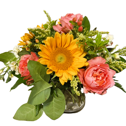 A vibrant floral arrangement featuring large yellow sunflowers, pink coral roses, solidago, peach hypericum, and green foliage in a small clear glass vase.
