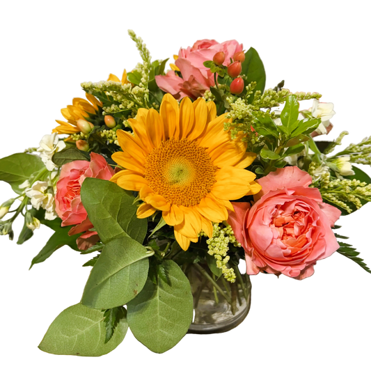 A vibrant floral arrangement featuring large yellow sunflowers, pink coral roses, solidago, peach hypericum, and green foliage in a small clear glass vase.