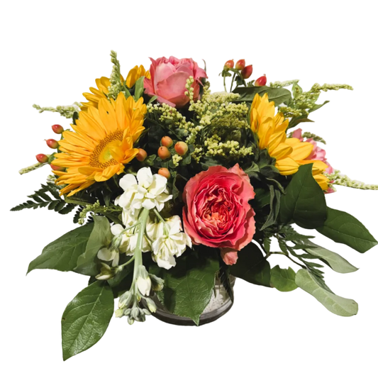 A vibrant flower arrangement in a small clear glass vase, featuring yellow sunflowers, pink roses, cream stock, and green foliage on a solid white background.