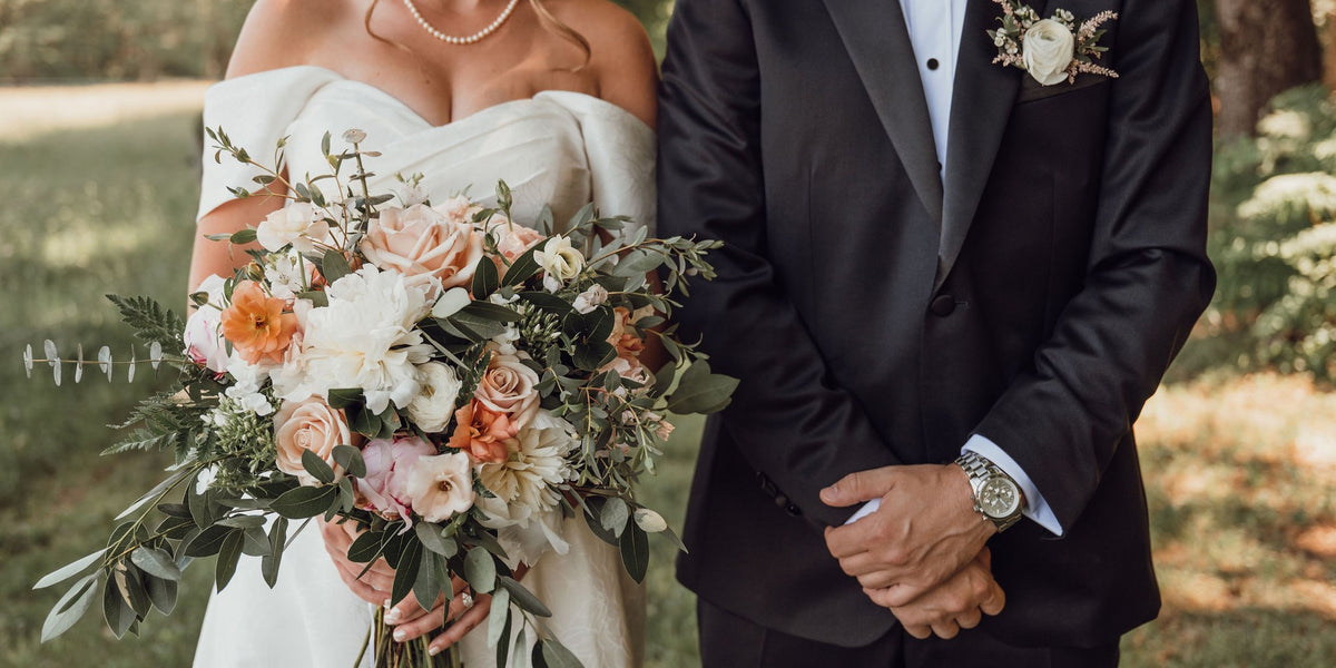 Couple in wedding attire with a bouquet of white, pink, and peach flowers outdoors.
