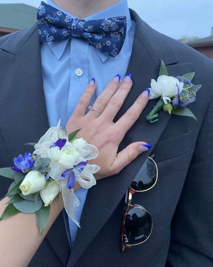 Person wearing a suit with a floral bow tie and ranunculus boutonniere.  A female hand with a white and blue  corsage rests on the lapel. 